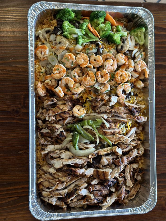 A foil catering tray of rice topped with shrimp, grilled chicken, steak, peppers and broccoli