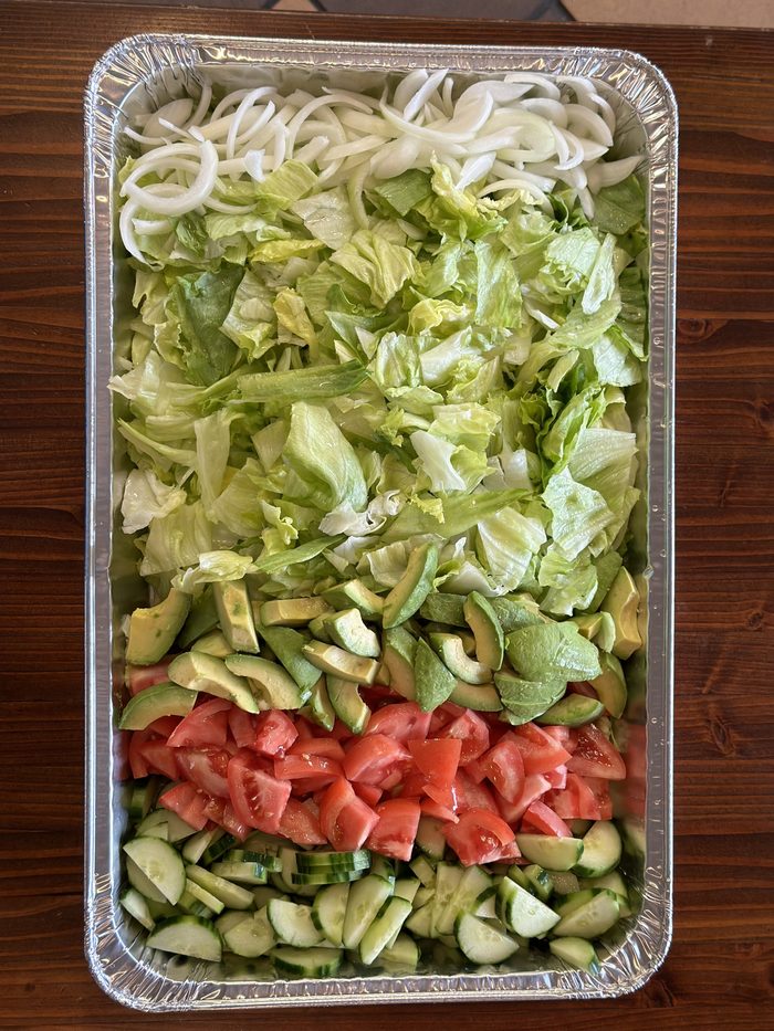 A foil catering tray of salad — onion, iceberg lettuce, avocado, tomato and cucumber in rows